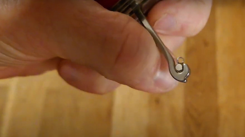 A close-up of a man's hand holding a Swiss Army Knife with the multi-purpose hook extended. Inside the hook is a small magnet.