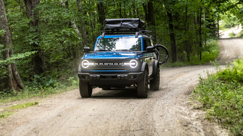 A blue Ford Bronco seen from front, driving on a forest road.