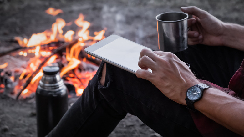 person using a tablet and drinking coffee by a campfire