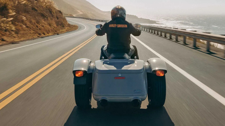 A man in a Harley-Davidson jacket riding along a mountain road on a 2024 Trike Freewheeler, back view