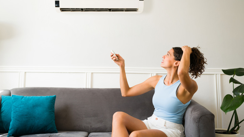 A distressed woman sitting on a couch pointing a remote control at a wall-mounted mini-split air conditioner.