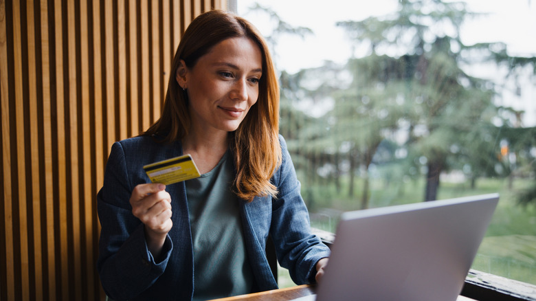 A woman holding her credit card in one hand as she looks at a laptop on a desk