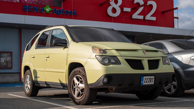A yellow Pontiac Aztek parked on pavement with its front to the road in front of a shop building.