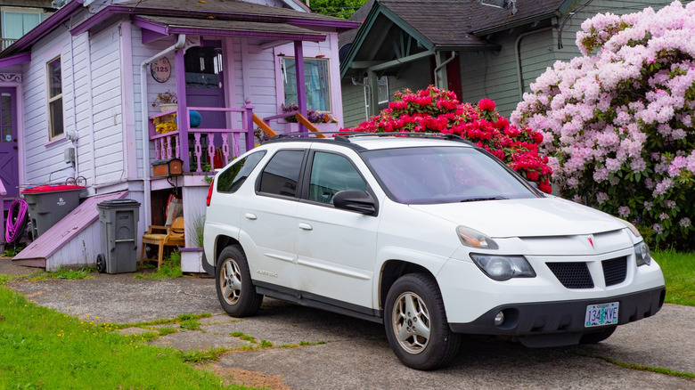 A white Pontiac Aztek standing in the driveway of a small, purple suburban house on a cloudy day.