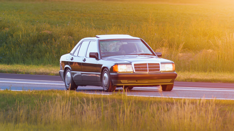 A 90s Mercedes sedan being driven through a rural setting with sun in the background.