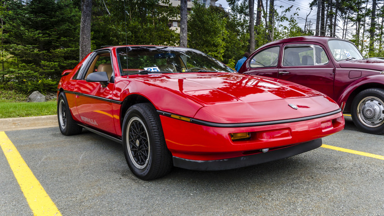 A 1980s red Pontiac Fiero parked near a VW beetle on an outdoor parking lot with trees in the background.