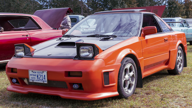 A tuned, red 1982 Toyota MR-2 sports car parked on green grass with other cars around it.