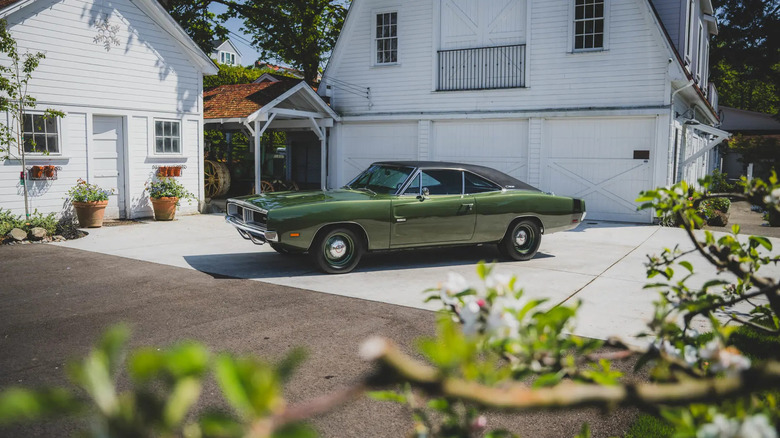 A dark metallic green 1969 Dodge Charger R/T parked in the driveway in front of an old white house.