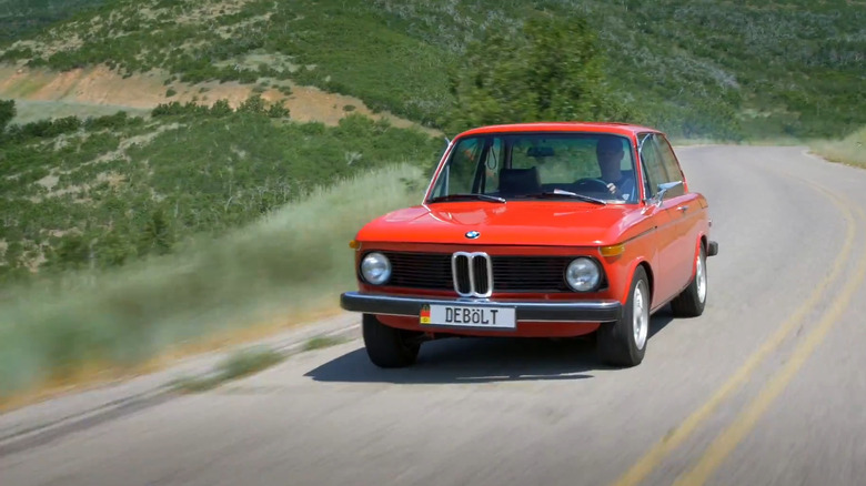 A red 1976 BMW 2002 Turbo car moving at speed down a curved mountain road with a valley in the back.