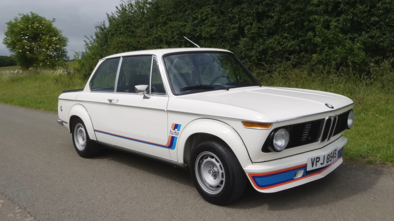 A cream colored BMW 2002 Turbo standing at the side of a narrow two-lane road at speed in the evening.