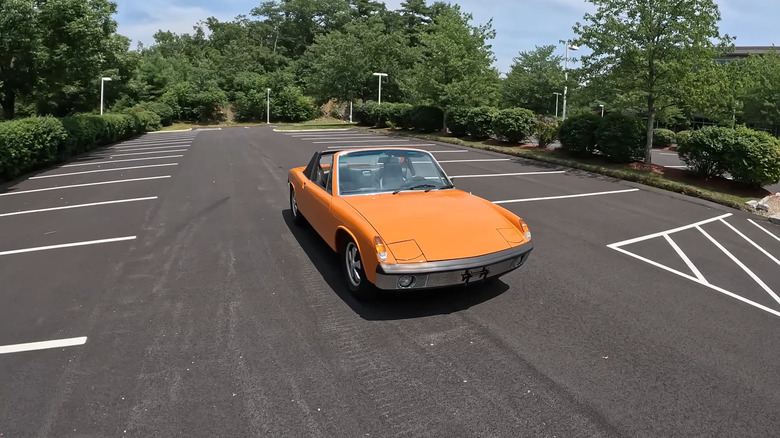 An orange Porsche 914/6 from 1970 standing in the middle of a completely empty parking lot with trees in the back.