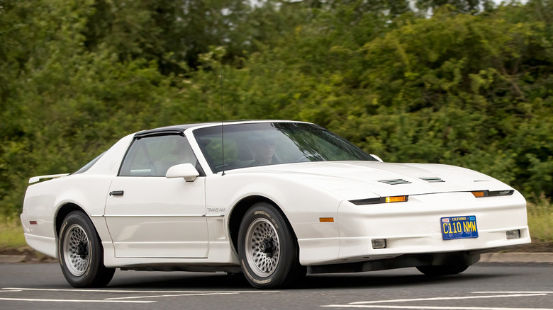 White 1986 Pontiac Trans Am driving along country road with brush in background, front-right 3/4 view