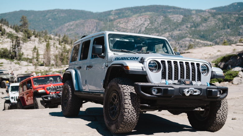 Three Jeep JL Wranglers driving up off-road trail in desert environment, front-right 3/4 view