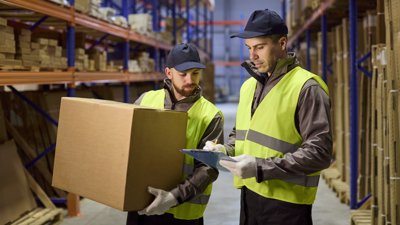 Man holding large box in a warehouse beside another man reviewing a clipboard.