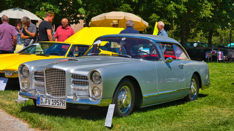 A grayish-blue Facel Vega HK500 luxury tourer being showcased at an outdoor event on a lawn next to other cars.