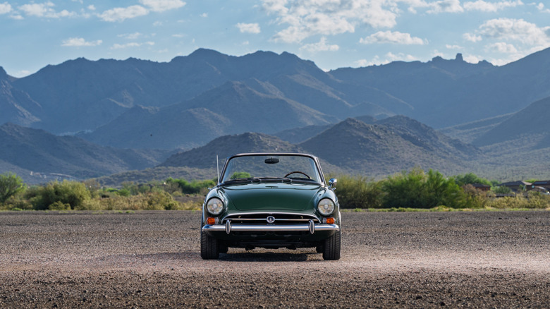 A dark green Sunbeam Tiger standing, front-facing the camera in front of some mountains in the distane.