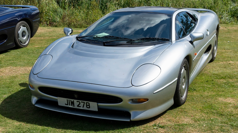 A silver 1980s Jaguar XJ220 parked on a grass field.