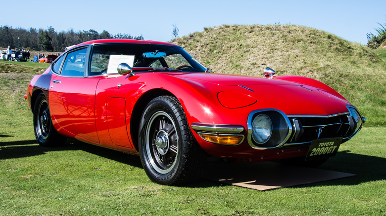 A red 1967 Toyota 2000GT parked on a grass field.