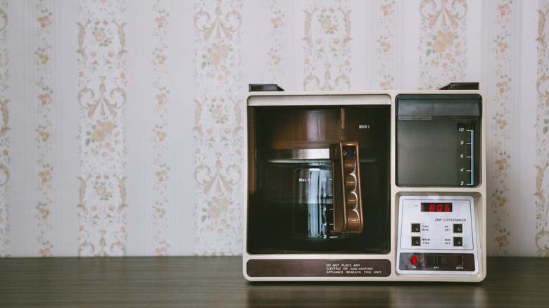 An old drip coffee machine sitting on a counter against a wallpapered wall