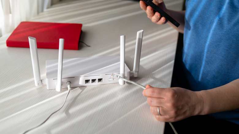 A person holding a phone in one hand and an Ethernet cable in another, next to a router and a red notebook on a white table