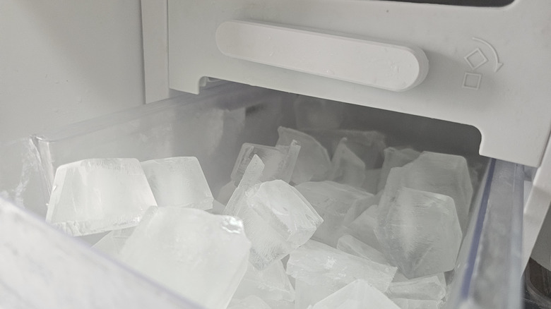 A refrigerator's ice maker compartment with a clear storage bin filled with ice cubes.