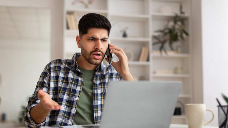 A man looking confused and frustrated while on the phone and sitting in front of his laptop