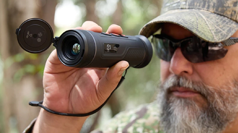 A bearded man in a camouflage hat looking through a black thermal vision monocular outdoors.
