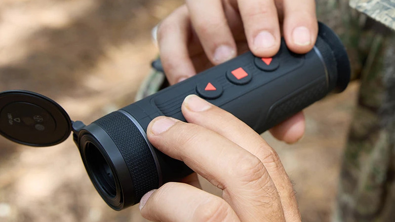 A pair of hands holding the ATN BlazeTrek Thermal Imaging Monocular with a blurred, bokeh effect backdrop.
