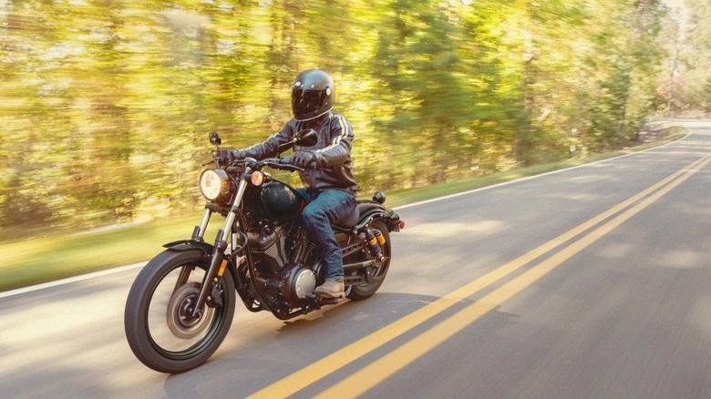 A rider in a full-face helmet riding a 2026 Yamaha Bolt R-Spec bike down a forested road with double yellow lines visible.