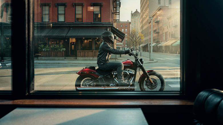 A male rider in full leather outfit and helmet on a red Harley Street Bob, photographed from inside a shop window moving by.