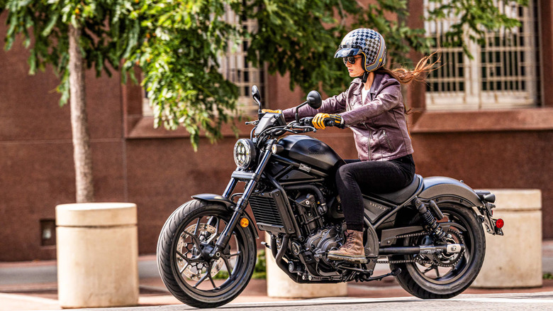 A woman rider in full gear and a helmet and boots riding a 2026 Honda Rebel 1100 through suburban streets.