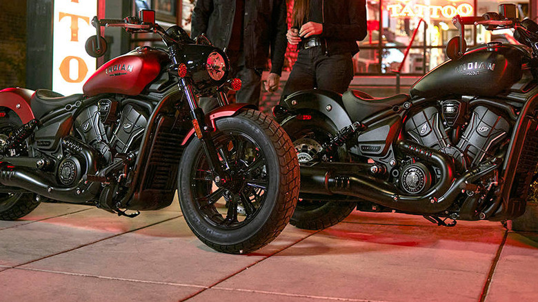 Two 2026 Indian Scout motorcycles -- one red and one black -- parked side by side in front of a café at night.