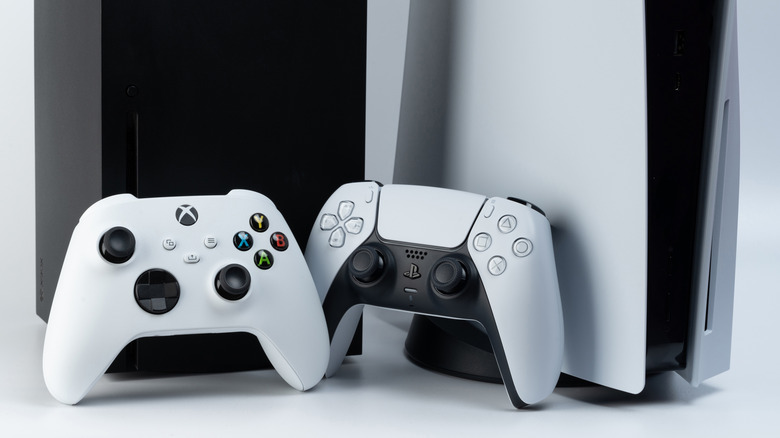 A close up of an Xbox Series X and PlayStation 5 with controllers in between them