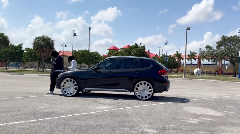 Two men in a parking lot, leaning against the hood of a parked BMW SUV with oversized chrome wheels.