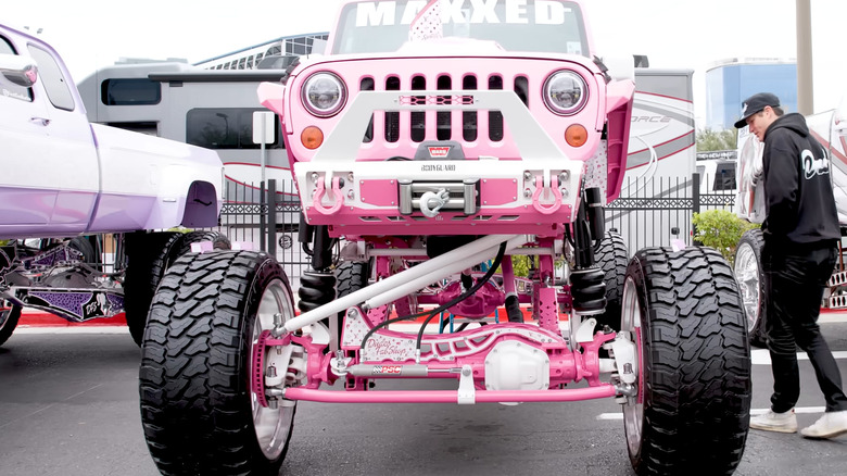 A man in all black clothing checking out a pink lifted Jeep that is parked at an outdoor auto show.