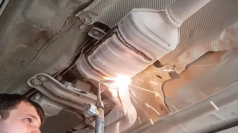 A man taking a blowtorch to the catalytic converter on the underside of a car on a lift jack.