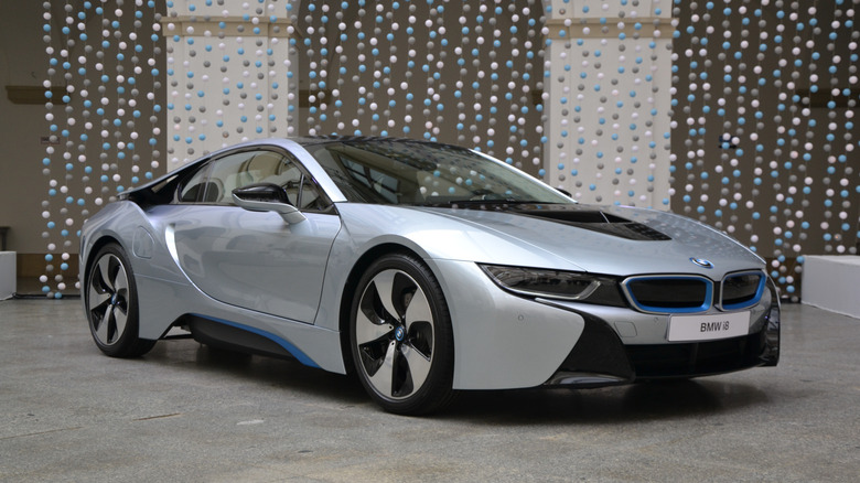 A silver and blue BMW i8 from the 2014 model year parked in the open on a paved surface.