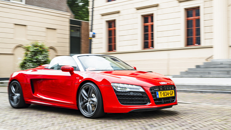 A first gen red Audi R8 driving on the street outside a beige building.