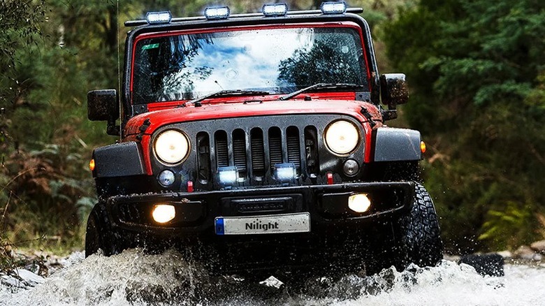 Red jeep with two light pods cruising through water.
