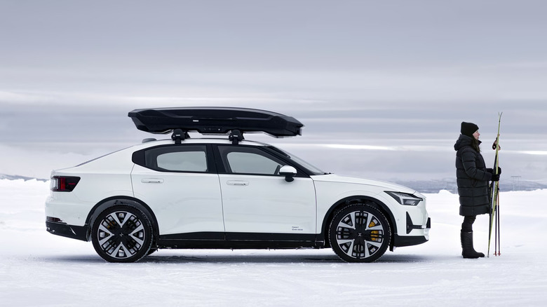 A white Polestar 2 with a Thule Motion 3 on its roof in a snowy area, person holding skiis upright next to it