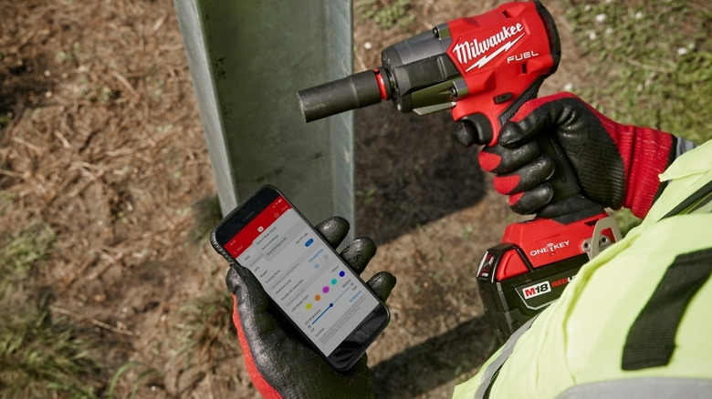 A person holding a Milwaukee Fuel Controlled Mid-Torque Impact Wrench and a phone with the One Key app.