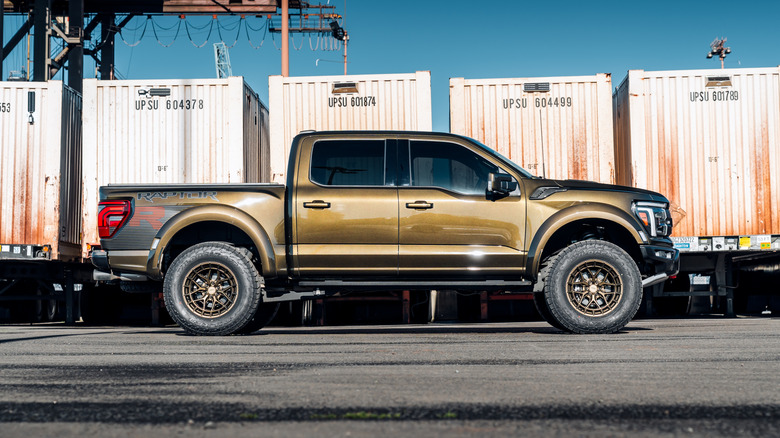A side-view of a chrome gold Ford F-150 Raptor parked near shipping containers in a dockyard.