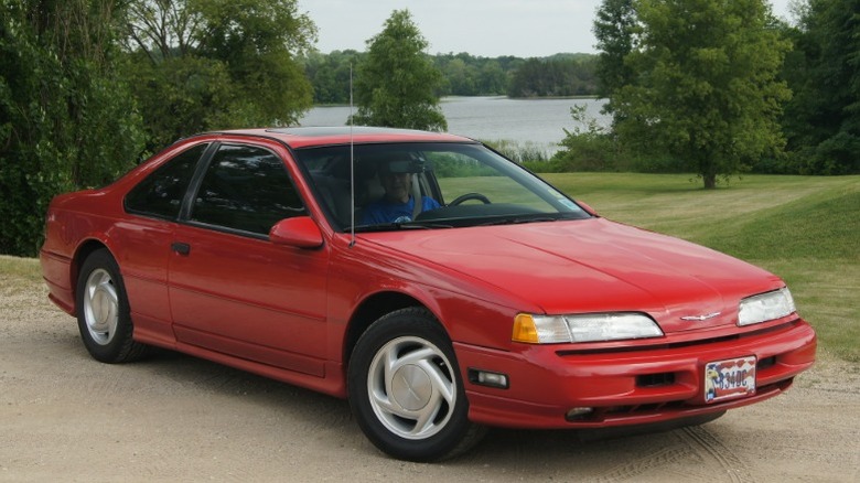Red Ford Thunderbird Super Coupe parked on dirt with greenery and lake in the background