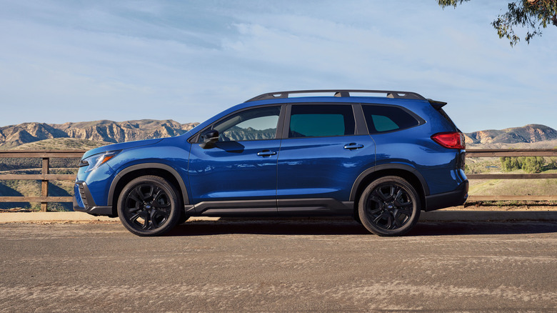 Side profile of a blue 2027 Subaru Ascent parked on a country road, mountains in background