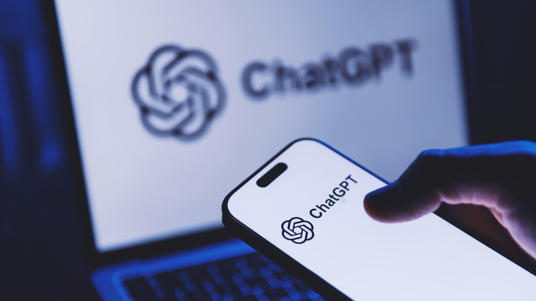 Person using ChatGPT on a phone with a blurred laptop in the background showing the ChatGPT logo