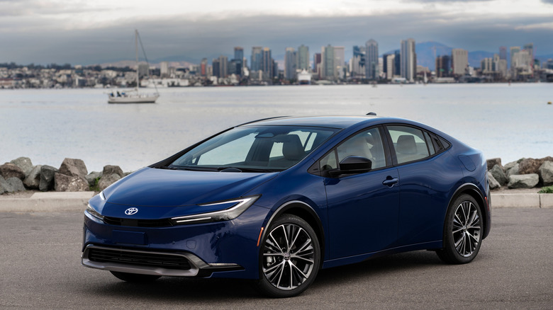 A blue 2026 Toyota Prius parked near water with a cityscape in the background