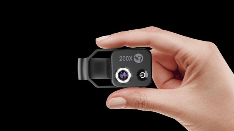 A person holding a Apexel 200X clip-on smartphone microscope lens against a black background