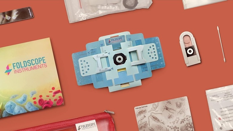 A Foldscope paper microscope kit on an orange surface, featuring the blue paper microscope, a red pouch, an instruction guide, and various accessories
