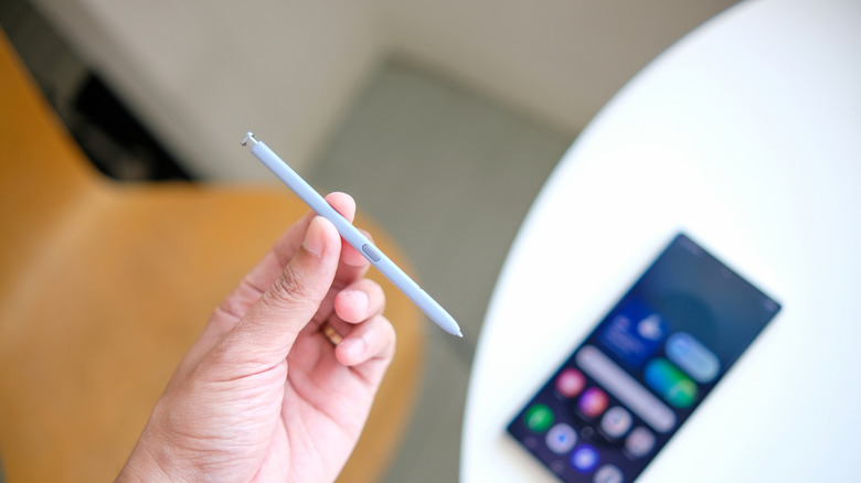 A hand holding up a stylus above a phone sitting on a white table
