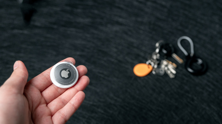 An AirTag held in someone's hand with keys in the background out of focus